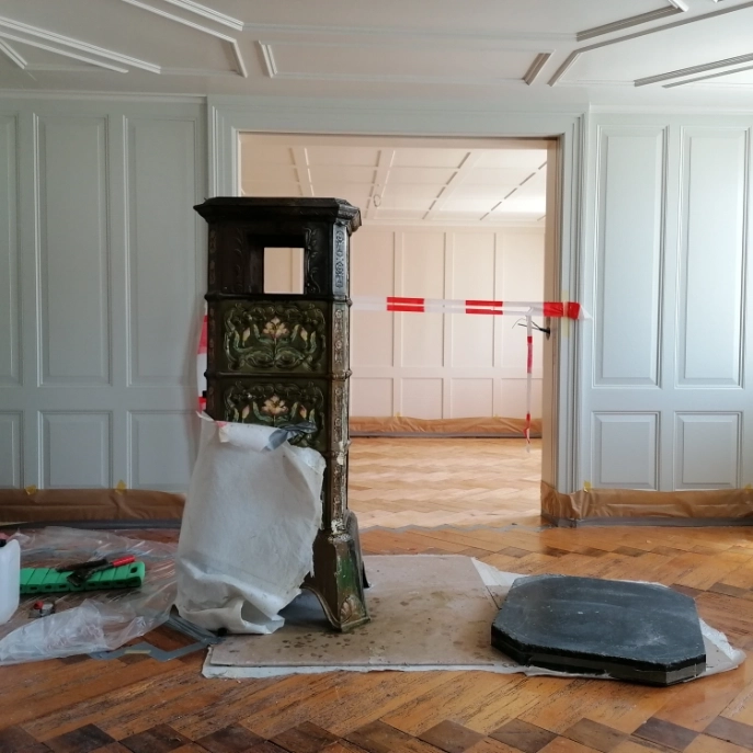 Raum mit weissem Täfer und Parkettboden, in der Mitte freistehender Ofen auf Folie