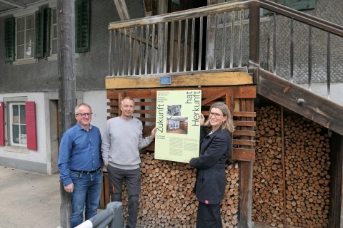 Gruppenfoto beim Eingang des Hauses vor der Holzbeige, Plakat wird präsentiert.