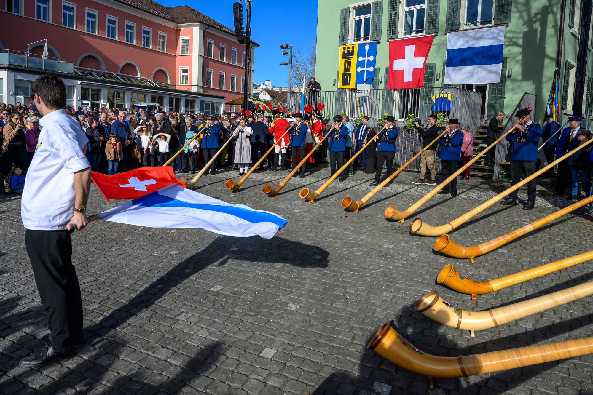 Fahnenschwinger mit der Zuger- und der Schweizerfahne, umringt von Alphornbläsern.